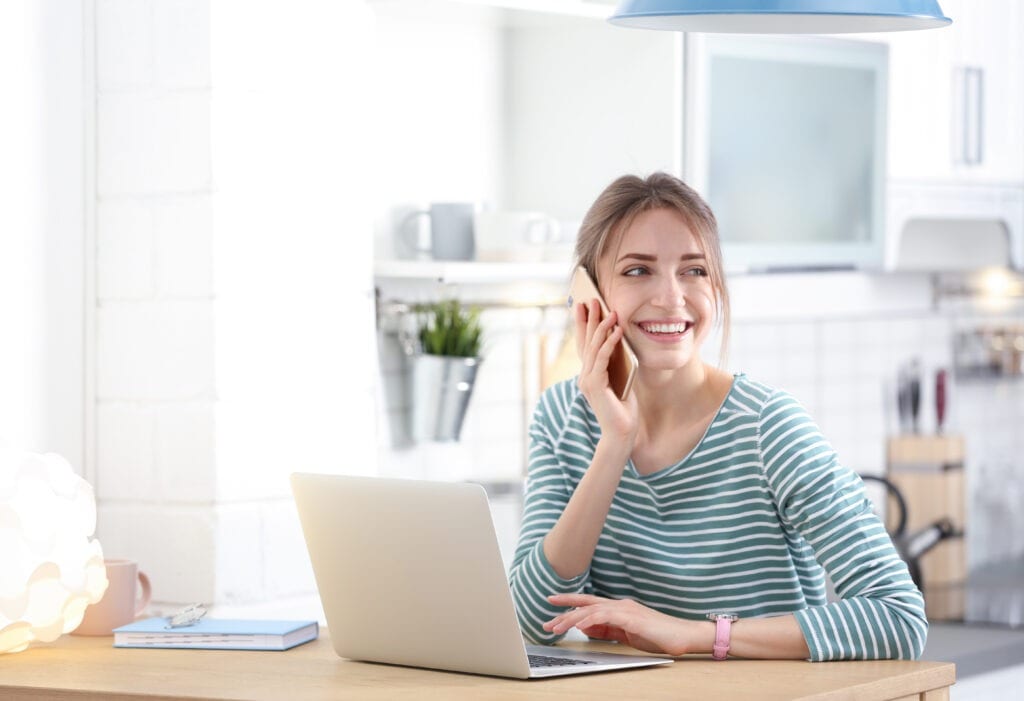 Happy young woman with laptop talking on phone at home