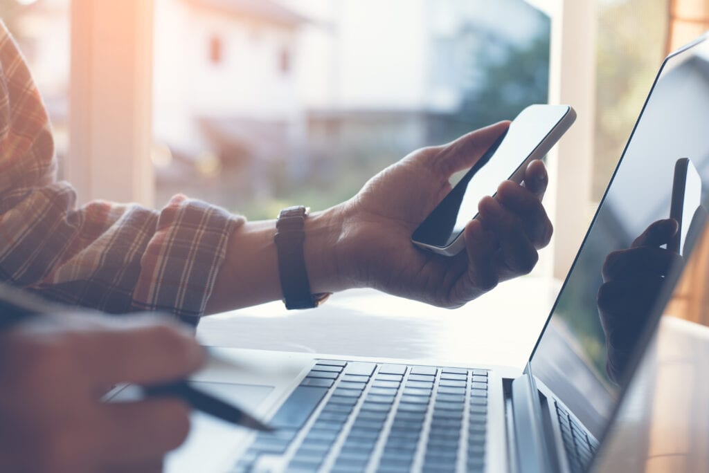 Close up of casual man or freelancer working on laptop computer and holding mobile smart phone with reflection on blank screen, working from home or casual business concept, vintage style.