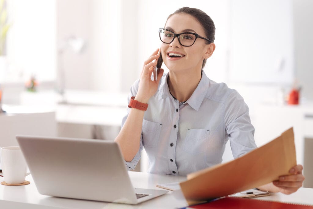 Express positivity. Young businesswoman holding phone near ear wearing glasses while being at workplace