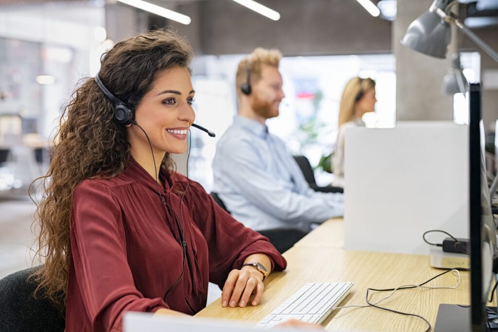 Service desk consultant talking on hands-free phone. Smiling call center operator with headset working on support hotline. Busy call center agents sitting in a row in a modern office, offering customer care service.